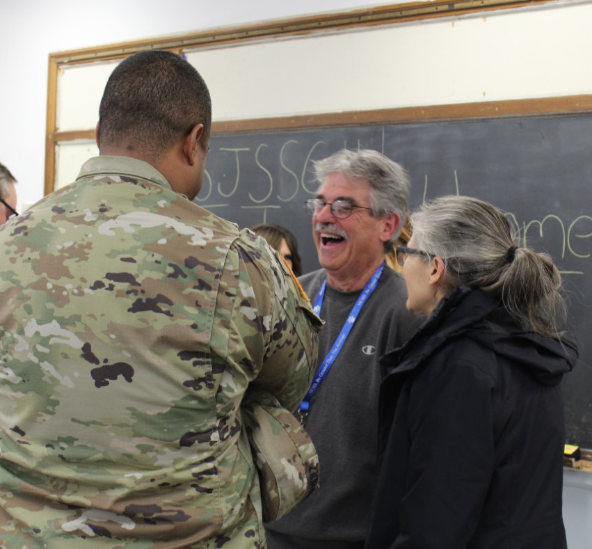 A man with light and dark gray hair wearing glasses laughs as he talks to another man and a woman.