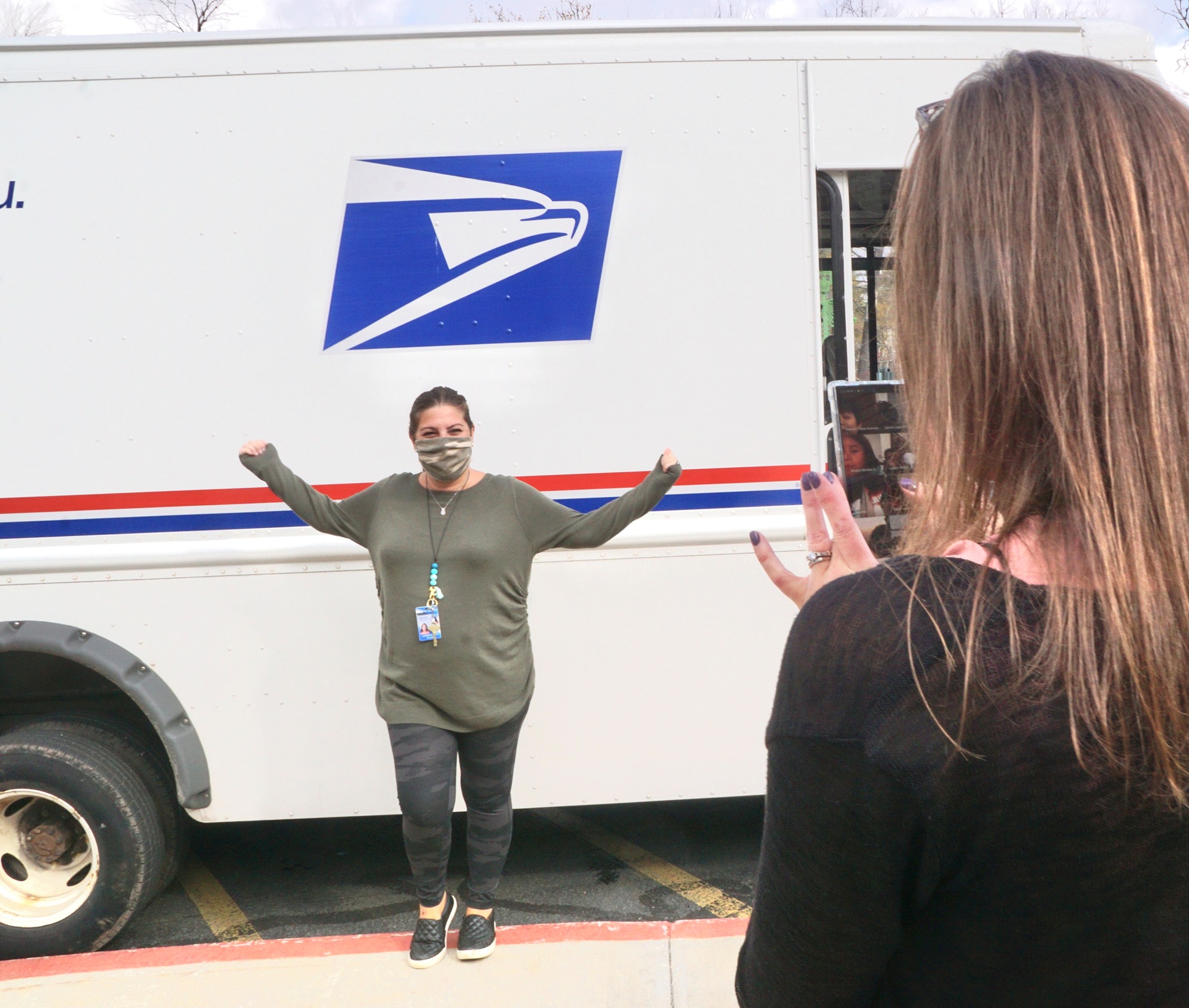 a teacher is filming a postal worker. The postal worker is standing beside a mail truck with her arms outstretched. 