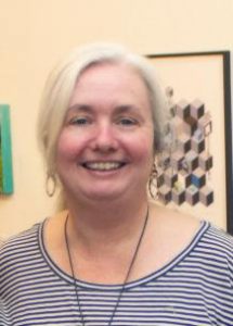 A woman smiles, has blonde hair pulled back in a ponytail, wearing a blue and white striped shirt, necklace and earrings