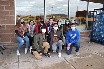 A group of nine high school students are in front of a supermarket, all wearing masks. Three are kneeling and six are seated behind them