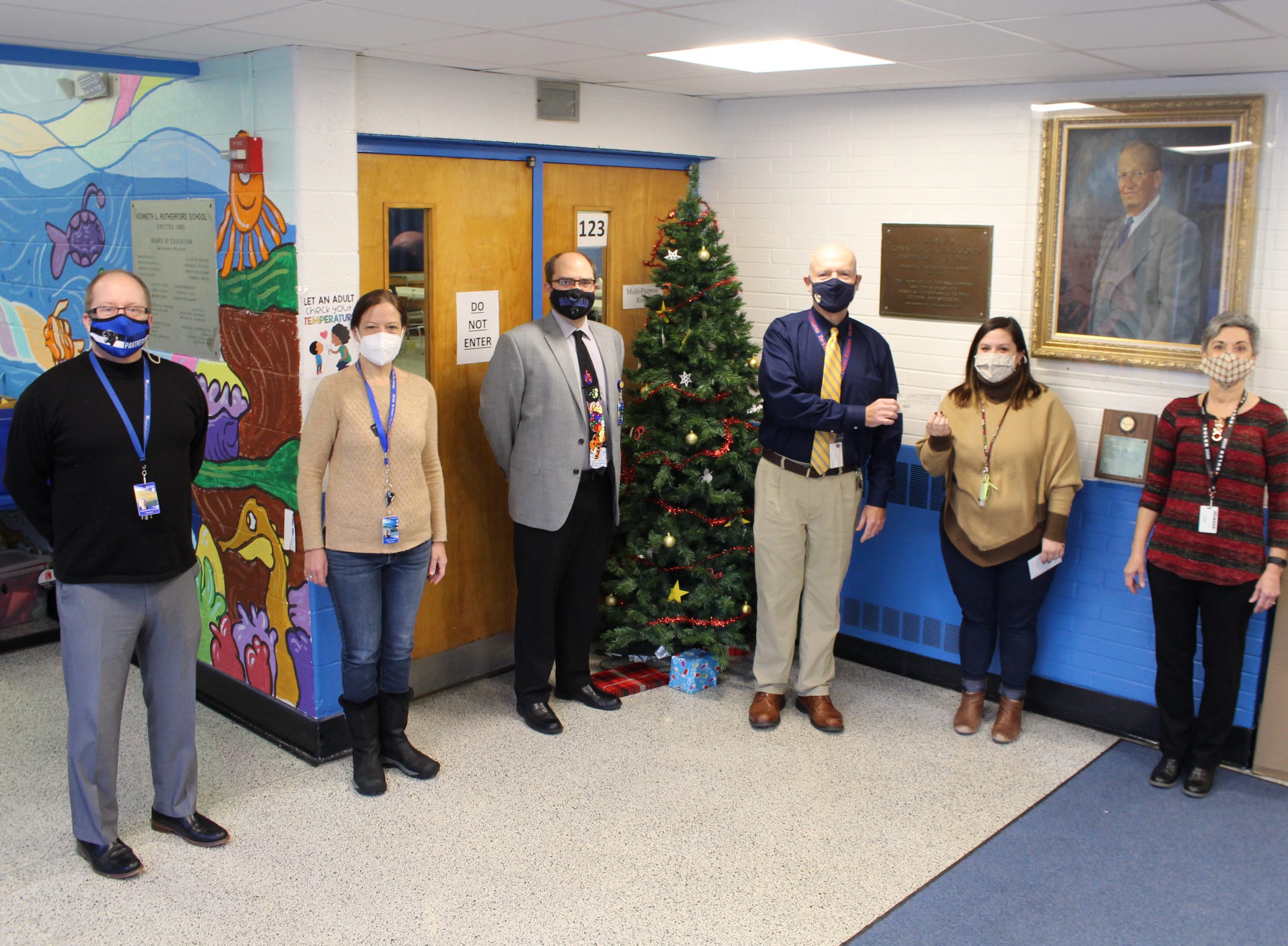 staff members are standing in the school hallway near a Christmas tree. Mike Williams is handing a check for $500 to Sarah Mootz to buy presents for students in need 