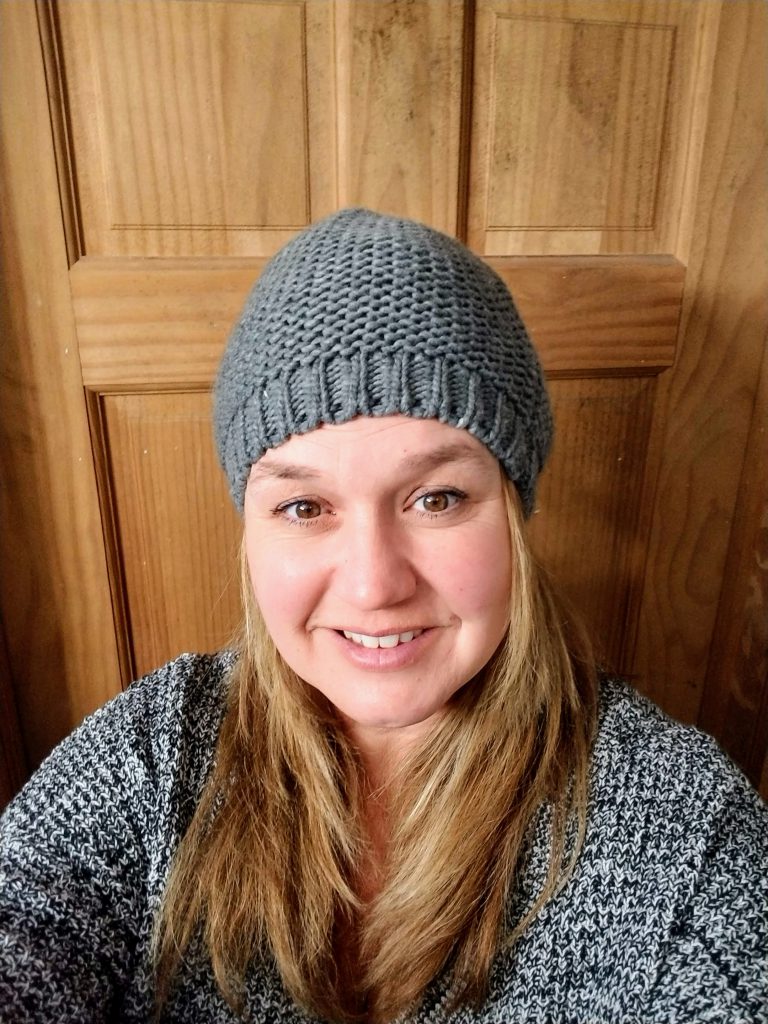 A woman with longer blonde hair, wearing a gray knit hat and sweater. She is smiling