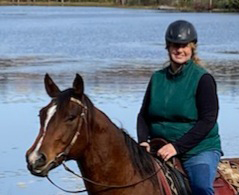 A woman wearing a riding helmet and green vest rides a horse. With water in the background.