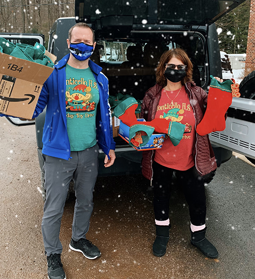 A man and a woman each wearing a Monticello High School Holiday Toy Drive shirt holds gifts and stockings outside of an open car back.