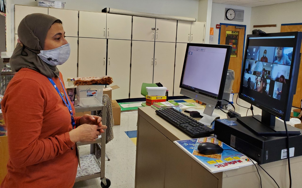 Ms. Elsayed is standing in front of a computer and speaking towards the computer. On the screen, you can see the images of students