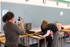 a reporter is holding a camera and filming a student working at a computer desk 