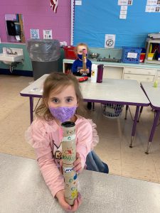 a young girl is seated at a desk holding a rain stick 