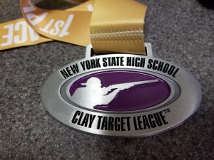A metal that says New york State High School Clay Target League. It has the profile of someone whooting a shotgun in the center and hangs on gold fabric.