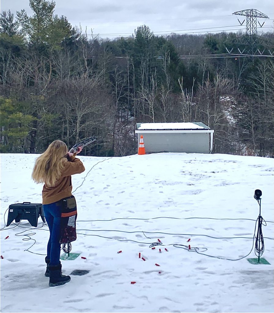 A young woman with long blonde hair shoots a target on a trap shooting field.