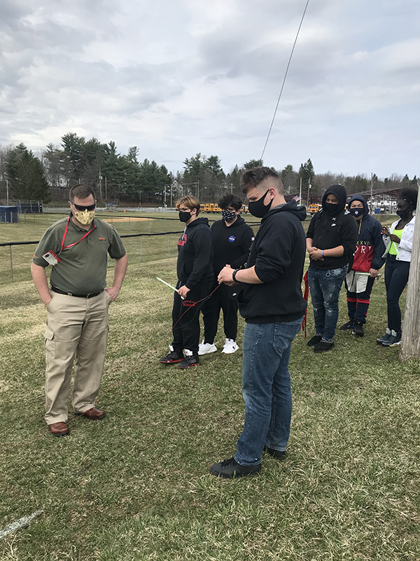 Outdoor scene, green grass cloudy sky. A group of high school students and a teacher stand with their rockets to launch.