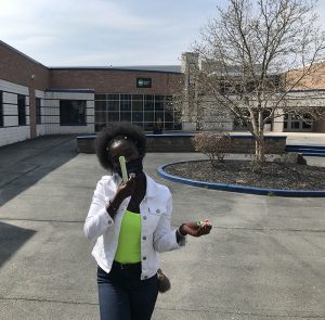 A high school young woman wearing a white jacket and bright green shirt holds up her green rocket.