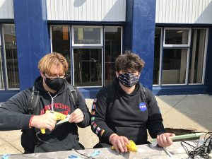 Two male high school students wearing black shirts and masks sit at a table making rockets out of green paper.