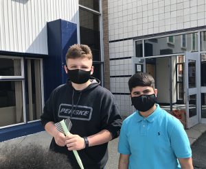 Two male high school students, one wearing a black t shirt and black mask and the other with a light blue shirt and black mask. The boy on the left is holding a green rocket made of paper.
