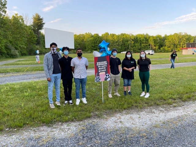 A group of six high school students standing by a sign that says Welcome to AOF Movie Night. There is a drive-in movie screen in the background.