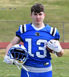 A high school senior boy with short brown hair is standing on a field of grass. He is wearing a blue football uniform with the  number 13 in white. He is holding a footballhelmet in his right hand and a football in his left.