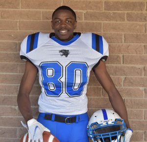 A high school senior boy wearing a football uniform, white jersey with blue number 80 and blue pants. He is smiling, has very short dark hair and is holding a football in his right hand and a helmet in his left. There is a brick wall behind him. 