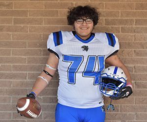 A high school senior boy in front of a brick wall. He is smiling, wearing glasses. He has a short crop of curly hair and is wearing a white football jersey with the number 74. He is holding a football in his right hand and a helmet in his left.