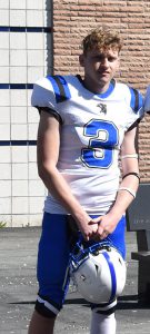 A high school senior boy wearing a football uniform stands with a brick building in the background. The jersey is white  with a blue number three and blue and black stripes on the shoulders. He is wearing blue pants and is holding a white football helmet with blue stripe.