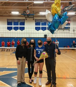 Four people, a woman in dark jacket and tan pants on the left, a young woman wearing black, then a high school senior wearing a volleyball uniform with the number three on it. She is holding flowers. On the right is a man dressed in dark clothes holding blue, gold and white balloons. All are wearing masks.