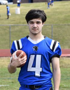 A high school senior boy with shorter dark hair. He is wearing a blue football jersey with the number 4 in white. He is holding a football with his right hand. There is grass in the background. 