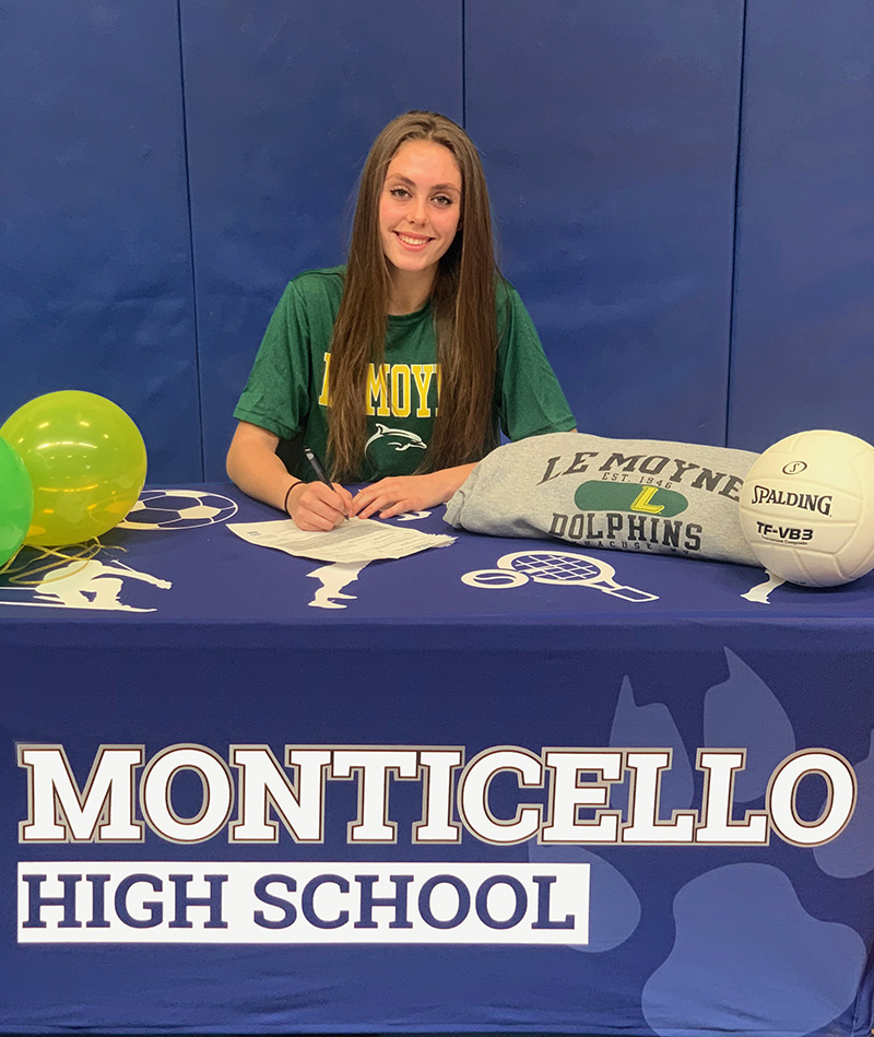 A young woman with long brown hair sits as a table which has a tablecloth that says Monticello High School. She is wearing a green t shirt with the college Le Moyne written on it. There are green and gold balloons and a volley ball on the table.