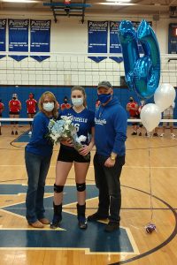 A woman on the left is wearing blue shirt and jeans and a white mask. In the center is a high school senior girl dressed in her volleyball uniform - black shorts and blue shirt. On right is a man wearing blue shirt and jeans. There are blue and white balloons next to him.