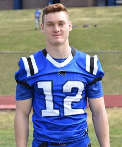 A high school senior boy with short light brown hair. He is standing on a grass field wearing a blue football uniform with the number 12 in white on it. There are white and black stripes on the shoulders.
