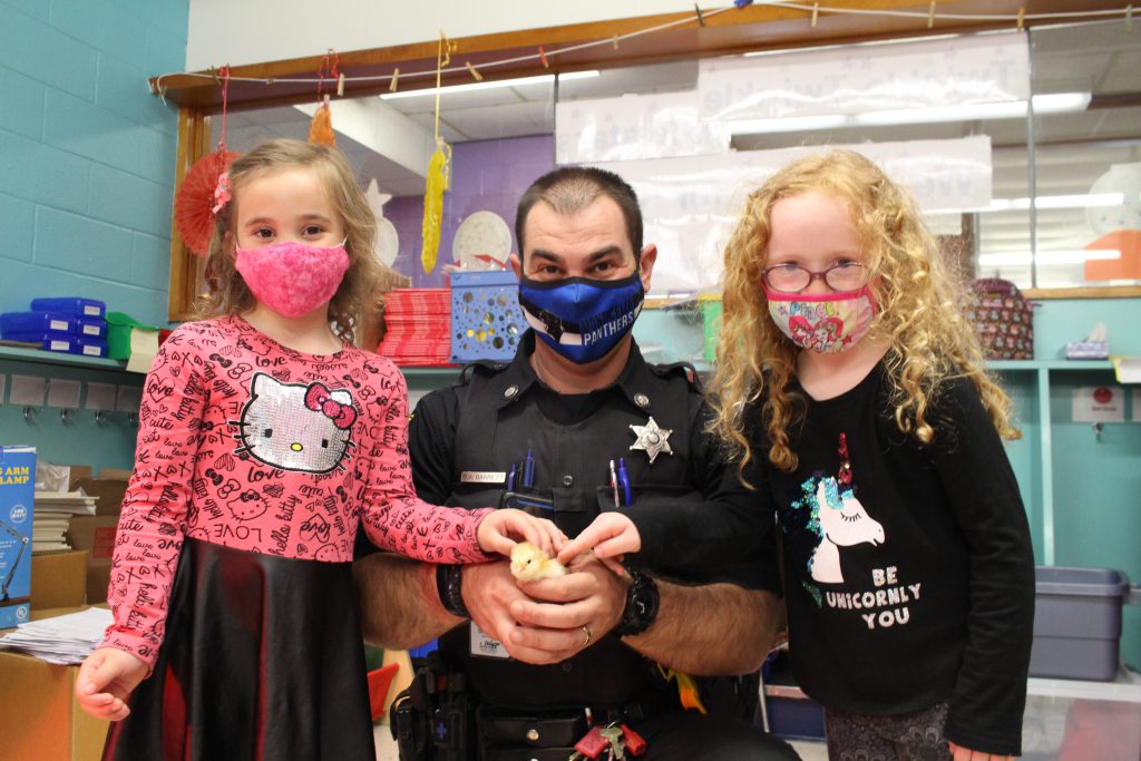 two young girls are standing with a police officer. The police officer is holding a yellow chick in his hand and the girls are petting it. 