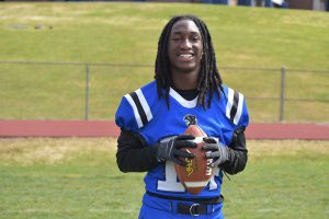 A high school senior boy with long braids smiling, wearing a blue football jersey with white stripes on the shoulders.