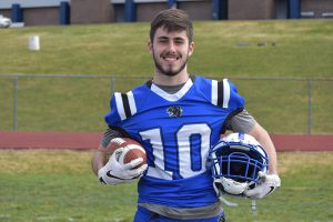 A high school senior boy  wearing a blue football jersey with the number 10 in white. He is holding a football in his right hand and a white football helmet in his left. There is green grass all around him.