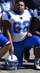 A high school senior boy kneeling on the ground. He is dressed in a football uniform, a white jersey with the number 63 in blue, and blue pants. He has his hand on a white helmet that sits on the ground.