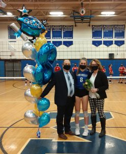 A man on the left wearing a suit and tie holds a large bunch of blue, gold and silver balloons. Next to him is a high school senior girl in a blue t shirt with the number 8 on it and black shirts. Next to her is a woman holding flowers, wearing a black jacket and plaid pants.