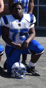 A high school senior football player, on one knee. He has short black hair. Uniform has a white  jersey with blue number 75 and blue pants. A white helmet with a blue stripe sits on the ground.