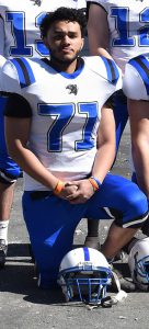 A high school senior boy wearing a fllotball uniform. He has darker short hair. The jersey is white with the number 71 in blue and blue pants. There is a white and blue football helmet in front of him on the ground. He is kneeling.