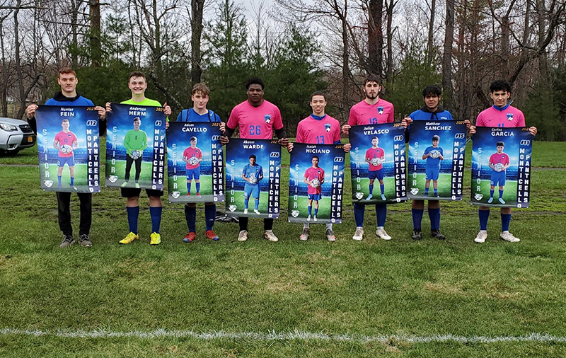 A photo of eight high school seniors, dressed in soccer uniforms, some green, some pink. Each is holding a large banner in front of them with their picture and number.