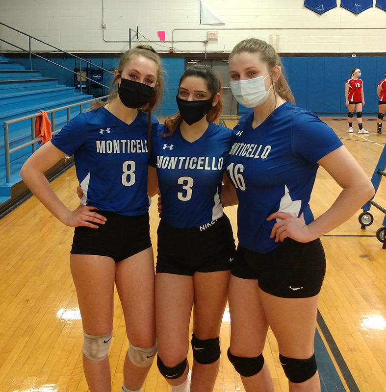 Three high school senior girls, all wearing masks. They are dressed in volleyball uniforms, black shorts and blue shirts that say Monticello.