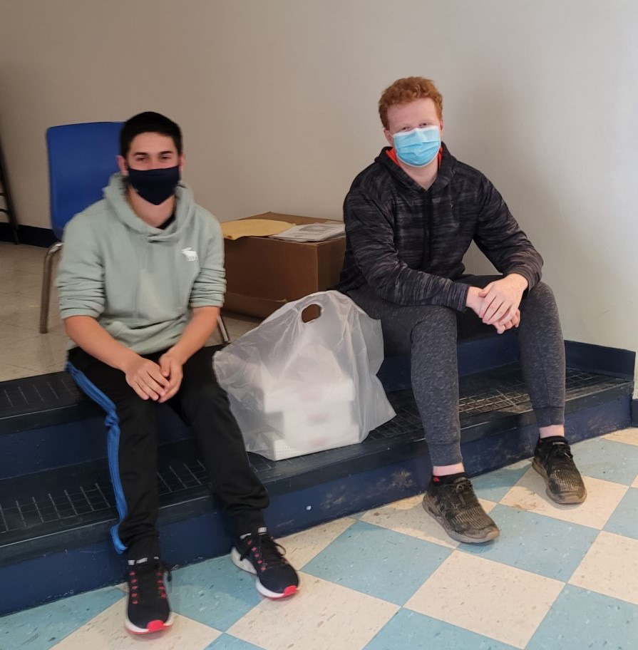 Two male high school students sit on a step inside the school. Between them is a bag with several boxes of donuts in it. 