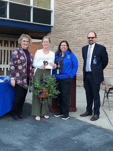 A woman wearing a white shirt and gray pants holds a hanging plant and certificate. She is standing with a woman on the left, a woman on the right and a man at far right.