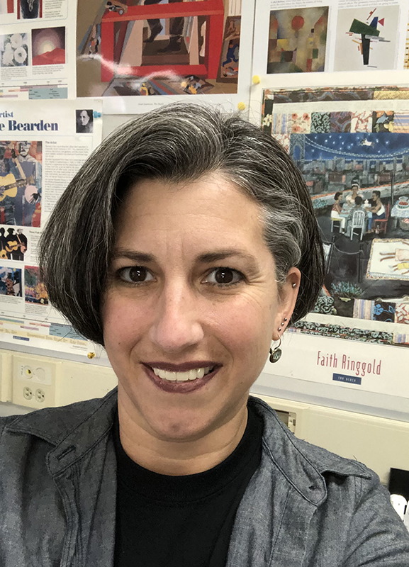 A woman with dark, shorter hair. She is smiling. Wearing a black shirt and black and white jacket