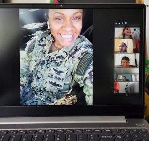 A female soldier, wearing army uniform is on a computer screen speaking with students