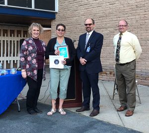 A woman wearing green pants holds a plaque and certificate. On the left is a woman and on the right are two men.