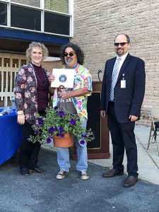 A man with long dark curly hair holds a plant and certificate. He is standing with a woman on the left and a man in a suit on the right.