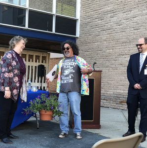 A man with long dark curly hair holds open his button down shirt to reveal a tshirt that says Now I have time to paint. A woman at left looks at it and laughs. A man on the right also laughs.