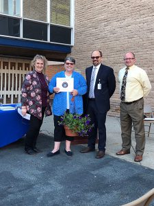 A woman wearing a blue jacket holds a plant and certificate. She is standing with a woman on the left, and two men on the right.