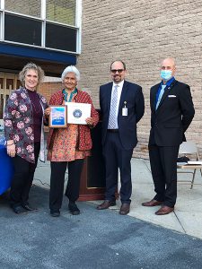 A woman with gray hair wearing a rust colored jacket holds two certificates. She is standing with a woman on the right and two men on the left, one has a mask on.