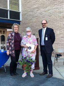 A woman in a flowered shirt, wearing a mask, holds a plant and certificate. There is a woman on the left and a man on the right.