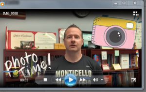 A man with short hair wearing a t shirt that says Monticello on it stands in front of a bookshelf with certificates on it. There is a graphic of a camera in the upper right corner and the words Photo Time! in the bottom left corner.