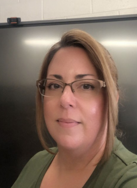 A woman wearing glasses and a green shirt with chin-length blonde-brown hair. The background is gray.