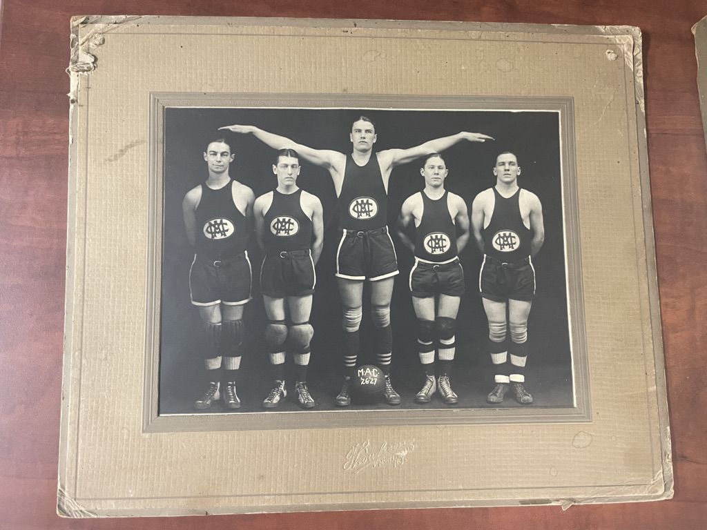 photo of five young men wearing basketball uniforms. There is a basketball at their feet that reads MAC 26-27. The photo is black and white and the uniforms appear to be from the 1920s. 