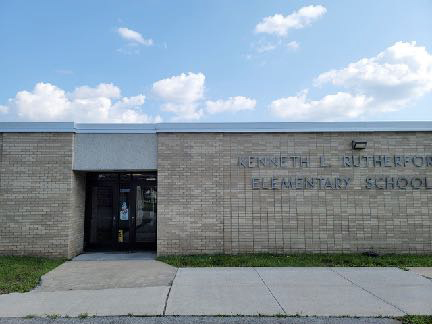 A tan brick build that says Kenneth L. Rutherford Elementary School. Doors are to the left.